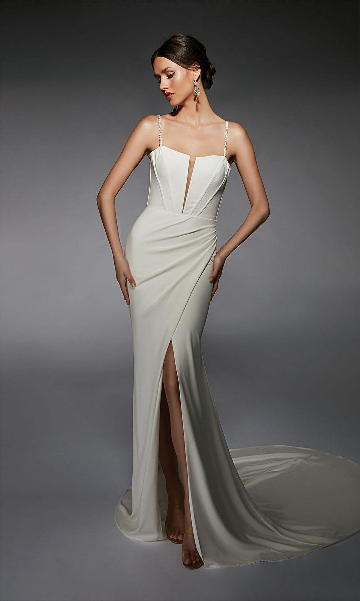 A woman poses in the Alyce 7104 long white dress, featuring thin straps, a deep V neckline, and a high slit, against a gray background.